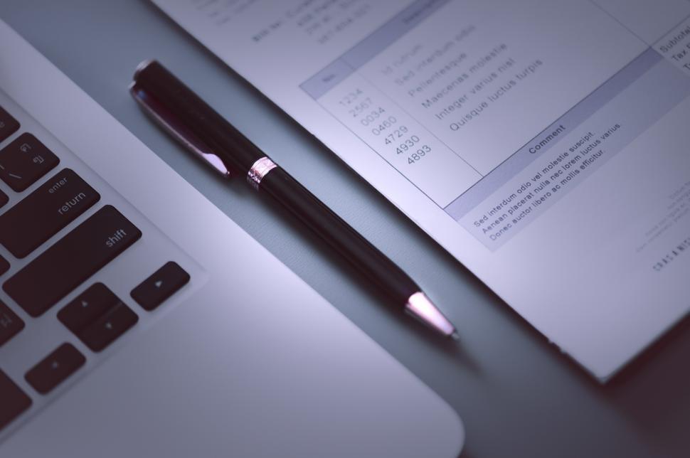 Closeup of a pen lying on a table, between the edge of a laptop and a sheet of paper.